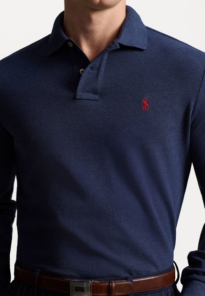 Marineblauwe polo met lange mouwen en een gestructureerde afwerking. Voorzien van een twee-knopen placket en een klein rood geborduurd logo op de borst. Bruine riem zichtbaar.