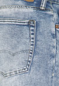 Vaqueros de mezclilla azul claro con un bolsillo trasero que tiene un diseño bordado, rematado con una etiqueta color crema y una costura amarilla en contraste.