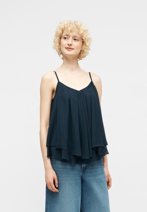 Kvinne med kort krøllete blondt hår iført en lagdelt mørkeblå ermeløs topp og lyseblå vide jeans mot en ensfarget bakgrunn.