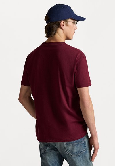Camiseta de algodón burdeos con mangas cortas y escote redondo, vista desde atrás. Acompañada de jeans de mezclilla azul claro y una gorra azul marino.