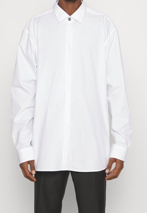 Hombre que lleva una camisa blanca de manga larga con botones y cuello clásico, combinada con pantalones negros, posando frente a un fondo liso.