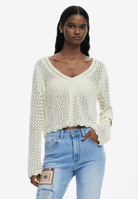 Cremfarbener, gestrickter Pullover mit V-Ausschnitt, offenem Maschenmuster, cropped Länge und gewelltem Saum. Kombiniert mit hellblauen, verwaschenen Jeans.