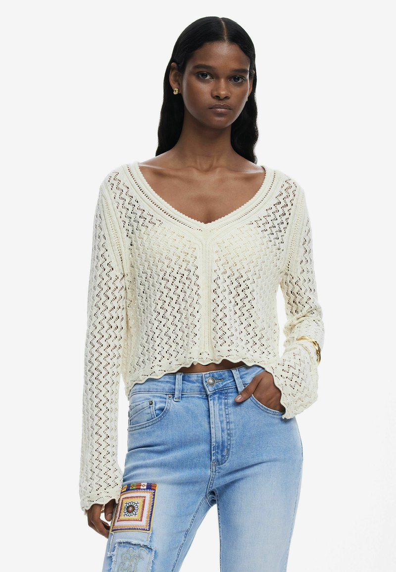 Cremfarbener, gestrickter Pullover mit V-Ausschnitt, offenem Maschenmuster, cropped Länge und gewelltem Saum. Kombiniert mit hellblauen, verwaschenen Jeans.