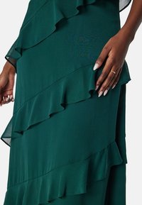 Zelené šaty s volánky a vrstveným lemem. Tkanina vypadá lehce a má jemný lesk. Dlouhé rukávy a hladká textura zvýrazňují design.