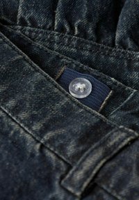 Temna jeansa tkanina, ki vsebuje žep z mornarsko modro nalepko. Jasna plastična gumb z obliko cveta je vidno postavljen na tkanini.