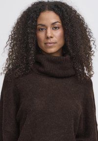 Maglione spesso di un marrone scuro con un ampio colletto a dolcevita ripiegato e una vestibilità larga. La trama appare morbida e accogliente, con una leggera peluria.