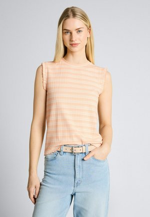 Donna bionda che indossa un top senza maniche a quadri color pesca infilato in jeans azzurri con cintura beige, in piedi davanti a uno sfondo semplice.