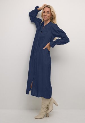 Abito lungo blu navy con chiusura a bottoni frontale, dettagli arricciati e maniche lunghe, abbinato a stivali beige con tacco.