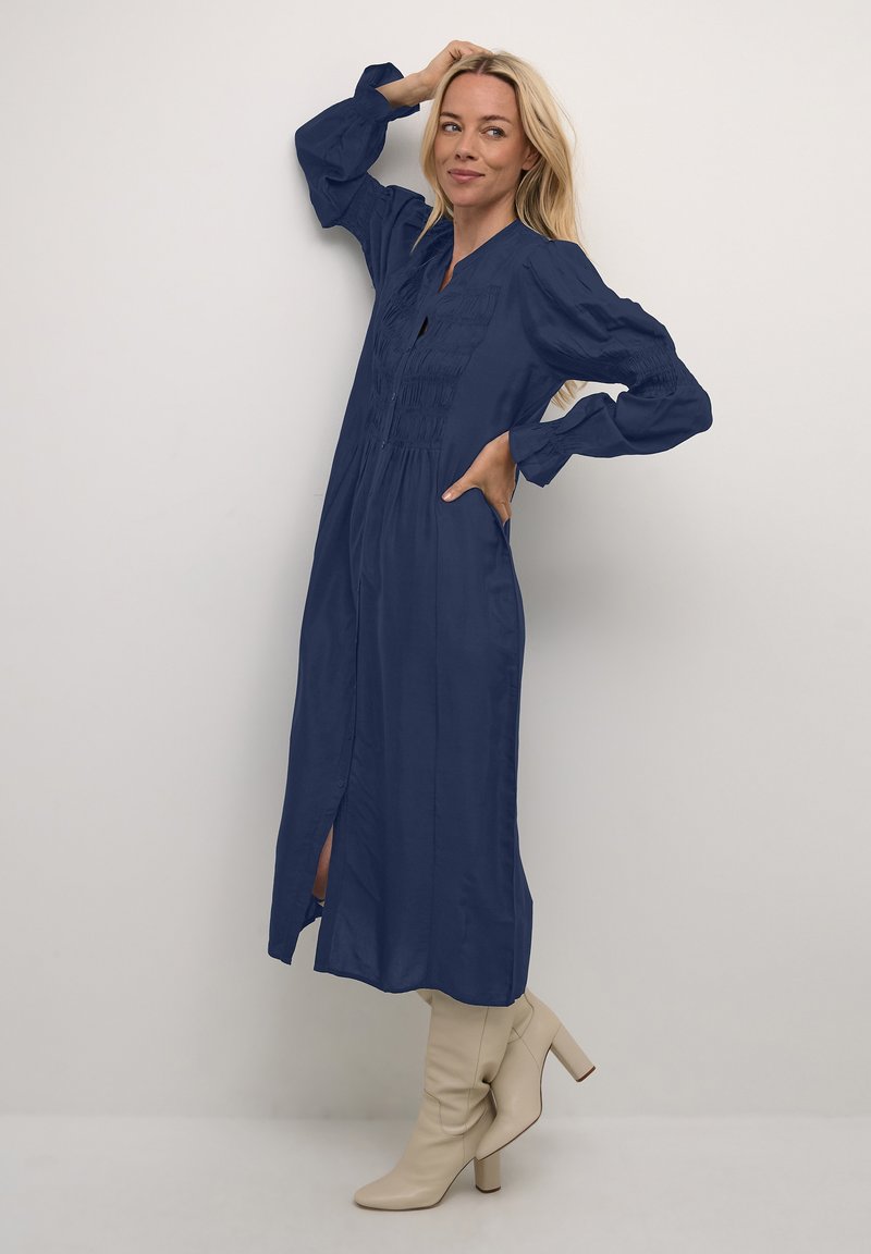 Abito lungo blu navy con chiusura a bottoni frontale, dettagli arricciati e maniche lunghe, abbinato a stivali beige con tacco.