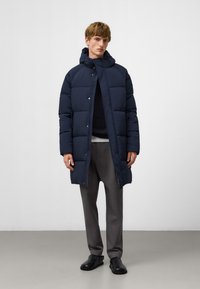 Granatowa kurtka puchowa z kapturem, napami, pikowanym wzorem i długimi rękawami. W komplecie z szarymi spodniami i czarnymi butami.