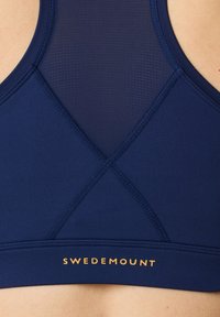 Granatowy biustonosz sportowy z teksturowanymi siateczkowymi panelami, krzyżowym designem na plecach oraz złotym logo "SWEDEMOUNT" na dole. Gładki, elastyczny materiał.