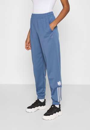 Osoba nosící modré tepláky Adidas s bílými pruhy a logy, v kombinaci s černobílými masivními teniskami.