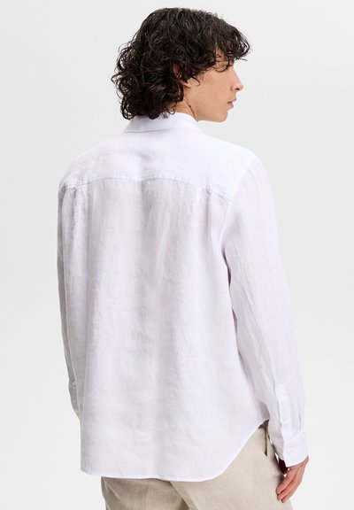 Jeune personne aux cheveux bouclés et foncés portant une chemise blanche en lin à manches longues et un pantalon beige, légèrement tournée de côté par rapport à la caméra.
