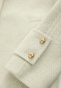 Crème blazer met een herringbone-textuur, voorzien van een mouw met twee gouden decoratieve knopen en een paspelzak aan de voorkant.
