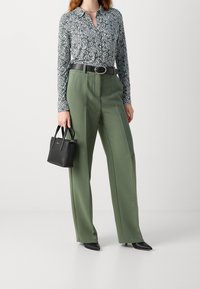 Groene op maat gemaakte broek gecombineerd met een zwart-wit gestippeld overhemd met knopen, gegord met een zwarte riem, en een kleine zwarte handtas.