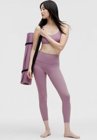 Jasnofioletowy zestaw sportowy składający się z dopasowanego topu z krótkim rękawem i legginsów z wysokim stanem, zestawiony z zwinięta matą do jogi zabezpieczoną czarnymi paskami.