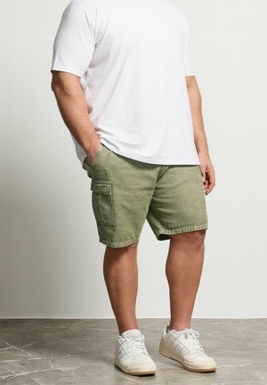 Mand iført hvid T-shirt, grønne cargoshorts med sidelommer og hvide sneakers, stående med hænderne i lommerne på gråt gulv.