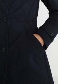 Námořnická bunda s texturovanou látkou, zdůrazněnou staženým pasem, bočními kapsami a knoflíkovými manžetami. Minimalistický design s funkčními detaily.