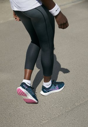 Chaussure de sport avec un design noir, bleu et rose, tige en mesh texturé et semelle intermédiaire à motifs, portée avec des leggings foncés et des chaussettes blanches.
