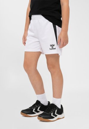 Barn iført hvide sportsshorts med sorte sidelister og logo, sorte sneakers med hvide chevroner, hvide sokker og en sort skjorte.