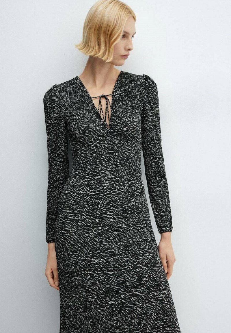 Abito nero con maniche lunghe e scollo a V. Caratterizzato da un tessuto testurizzato con un motivo a pois e un dettaglio con fiocco al collo. Design aderente.