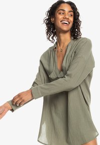 Blusa di colore verde oliva, ampia e comoda in un tessuto leggero e texturizzato, con scollatura a V profonda e maniche lunghe. Presenta un delicato dettaglio plissettato.