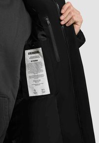 Svart jacka med en slät inredning. Har en ficka med dragkedja och en vit skötselöns som är fäst, med text och logotyper.