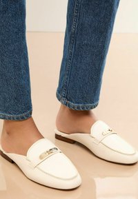 Witte leren instaploafer met een gouden accent, met een gladde textuur, ronde neus en lage houten hak, gecombineerd met blauwe jeans.