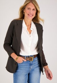 Donkerbruine getailleerde blazer met daaronder een frisse witte blouse, spijkerbroeken en een leren bruine riem. Glad materiaal met subtiele ontwerpen.