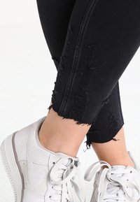 Black ripped jeans with frayed hems, paired with white sneakers. The jeans feature distressed detailing along the sides and a slim fit.