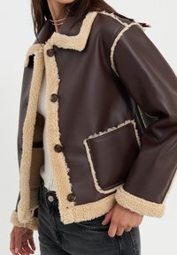 Brun kunstskinnjakke med beige shearling-fôr, knapplukking og en lommepatch. Har kontrastkanter og en avslappet passform.