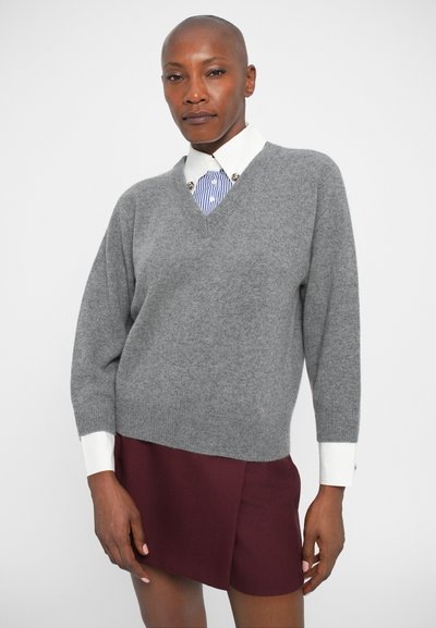 Grauer Woll-V-Ausschnitt-Pullover mit überschnittenen Schultern, über einem weißen Hemd mit blauen Streifen getragen, kombiniert mit einem bordeauxfarbenen Rock.
