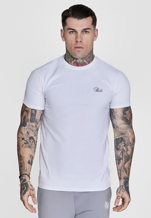 Muskuløs mann med omfattende tatoveringer på armer og nakke, iført en tettsittende hvit t-skjorte og grå bukser, vendt mot kamera med et nøytralt uttrykk.