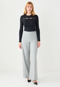 Schwarzes Langarmoberteil mit Cut-out-Design und graue, hoch taillierte, weit geschnittene Hosen, kombiniert mit schwarzen spitzen Schuhen. Glatte Texturen überall.
