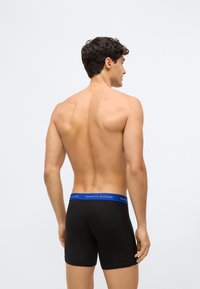Homem de torso nu visto de costas, vestindo boxers pretos com um cós azul da Tommy Hilfiger, contra um fundo claro e liso.