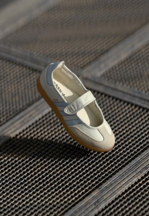 Chaussure Adidas Samba blanche avec bride velcro et semelle en gomme flottant au-dessus d'un fond de grille métallique texturée.