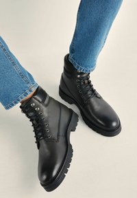 Czarne skórzane botki za kostkę z teksturowanym wykończeniem, okrągłym noskiem i sznurowanym przodem. Posiadają metalowe oczka oraz solidną podeszwę. Widoczne dżinsowe spodnie.