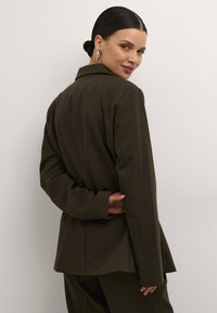 Mørkeolivenfarvet skræddersyet blazer med en struktureret silhuet, klippede slag og en enkelt rygslids. Glat tekstur, minimalistisk design.