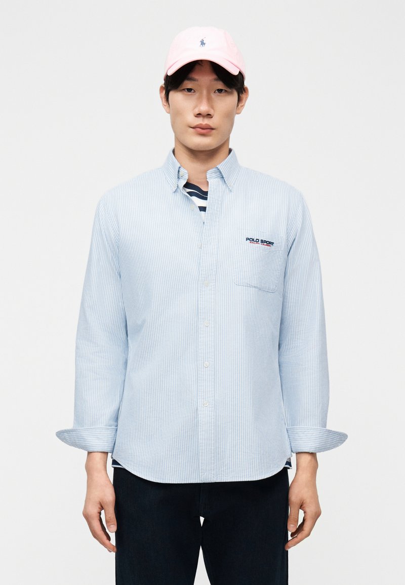 Νεαρός άνδρας που φοράει πουκάμισο με ανοιχτό γαλάζιο ριγέ και το λογότυπο "Polo Sport" μαζί με ροζ καπέλο, σε απλό λευκό φόντο.