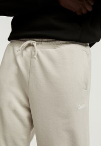 Beige Jogginghose mit Zugband in der Taille, seitlichen Taschen und einem kleinen weißen Logo am Oberschenkel. Hergestellt aus weichem, glattem Stoff.