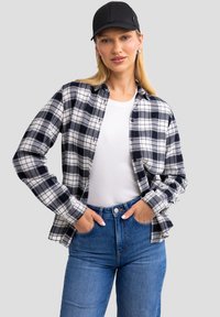 Schwarz-weiß karierter Flanellshirt über einem weißen, taillierten T-Shirt, kombiniert mit hochgeschnittenen blauen Jeans. Das Model trägt eine schwarze Mütze.