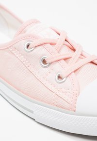 Zapatilla de lona rosa claro con puntera y suela de goma blanca, con ojales metálicos y cordones a juego. Detalles de tela texturizada visibles.