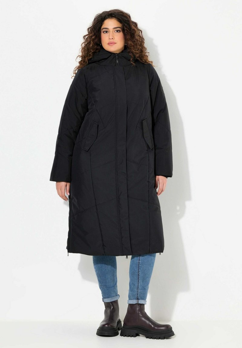 Svart vadderad jacka med huva, med en framdragkedja, två sidofickor och en midjelång design. Bärs över ljusblå jeans och mörka stövlar.