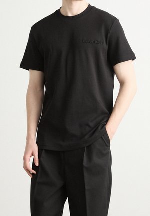 Mężczyzna noszący czarną koszulkę Calvin Klein z krótkim rękawem i czarne spodnie, stoi z jedną ręką w kieszeni na jednolitym tle.