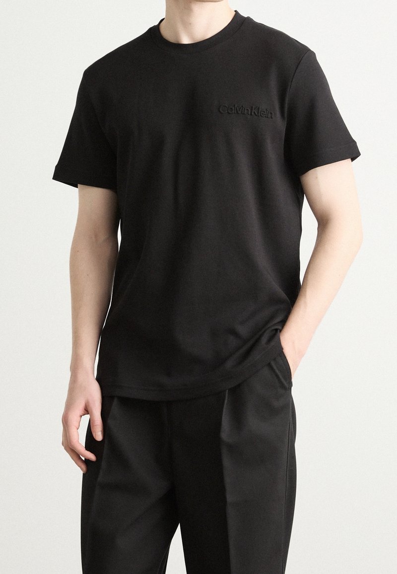 Homme portant un T-shirt Calvin Klein noir à manches courtes et un pantalon noir, debout avec une main dans la poche devant un fond uni.