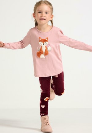 Nuori tyttö, jolla on palmikkohiukset ja yllään vaaleanpunainen pitkähihainen ketun paita, maroon-kasvikuvioiset leggingsit ja vaaleanpunaiset saappaat, seisoo yhdellä jalalla.