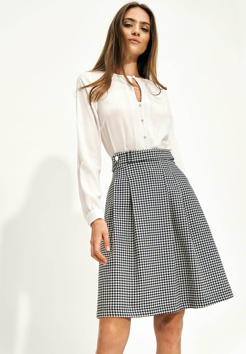 Kobieta ubrana w białą bluzkę z długim rękawem oraz czarno-białą spódnicę w kratkę do kolan na jednolitej tle.