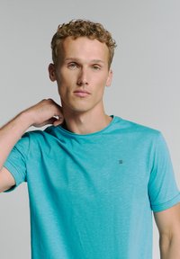 Turquoise t-shirt van een gladde stof met een ronde hals en korte mouwen, met een klein zwart logo op de borst.
