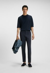 Granatowa koszula z krótkim rękawem z kieszonką, ciemne spodnie i ciemne buty wsuwane. Trzyma w ręku fakturowaną niebieską marynarkę z trzema guzikami.