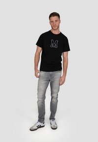 T-shirt noir à manches courtes avec un logo "M" brodé en blanc, porté avec un jean gris déchiré et des chaussures de sport noir et blanc.
